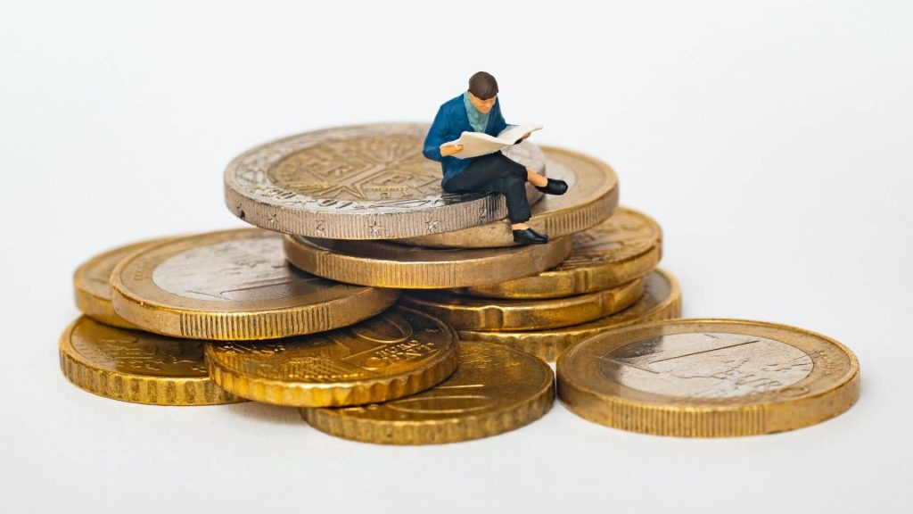 Small plastic figure of a man reading a newspaper atop a stack of Euro coins.